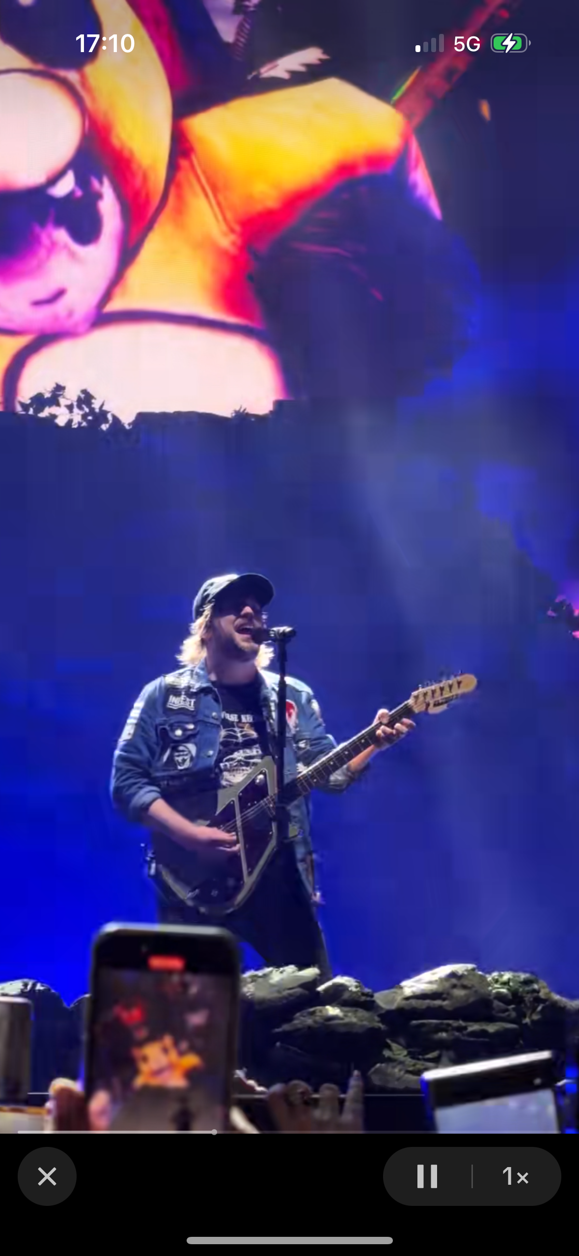 a male rock musician performs on stage with a guitar in hand, his microphone is visible as well.  the audience can be seen in the background, as well as bright lights and a large screen showing the image of a cat, possibly part of the band's set design.