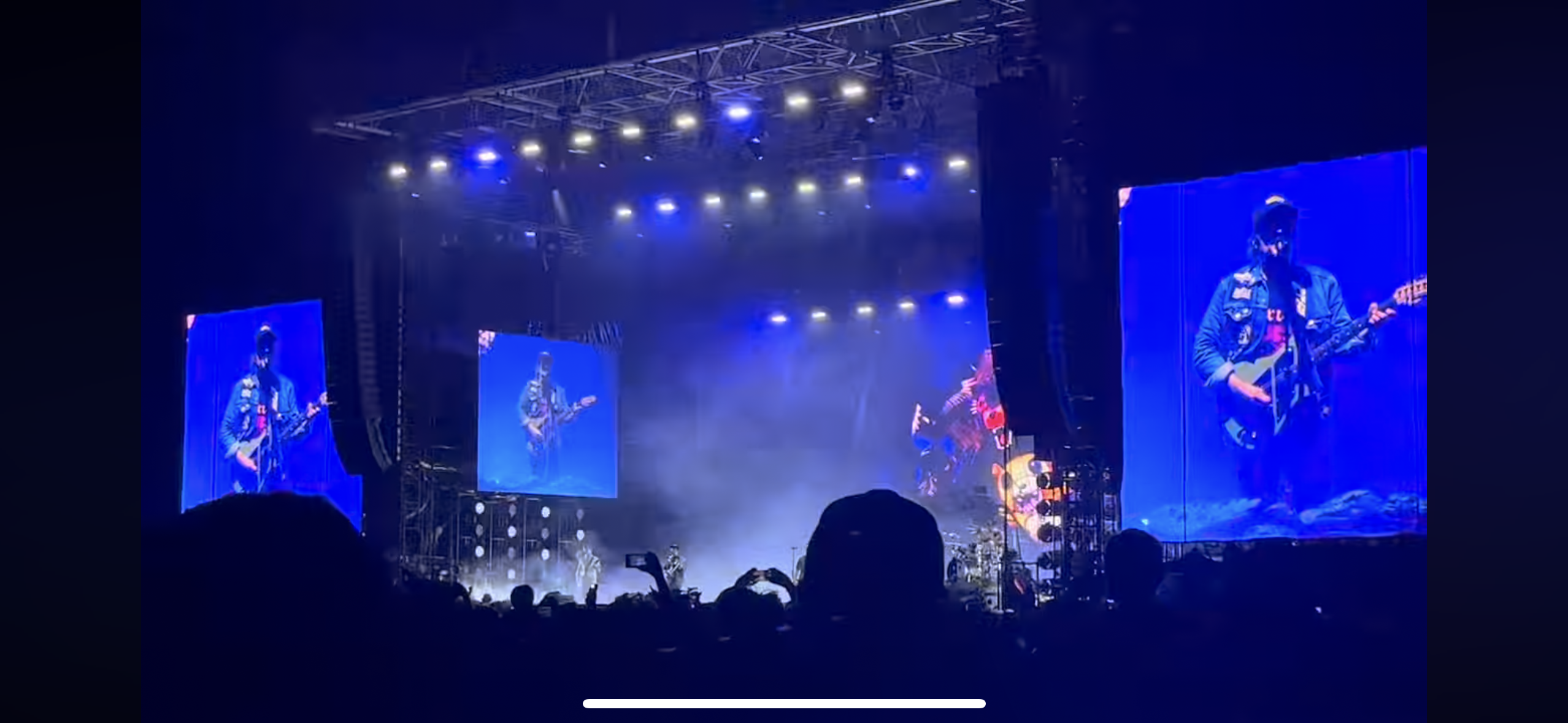 a concert stage with the band performing on a large stage with bright lights. the audience is seen in the foreground, silhouetted against the stage.