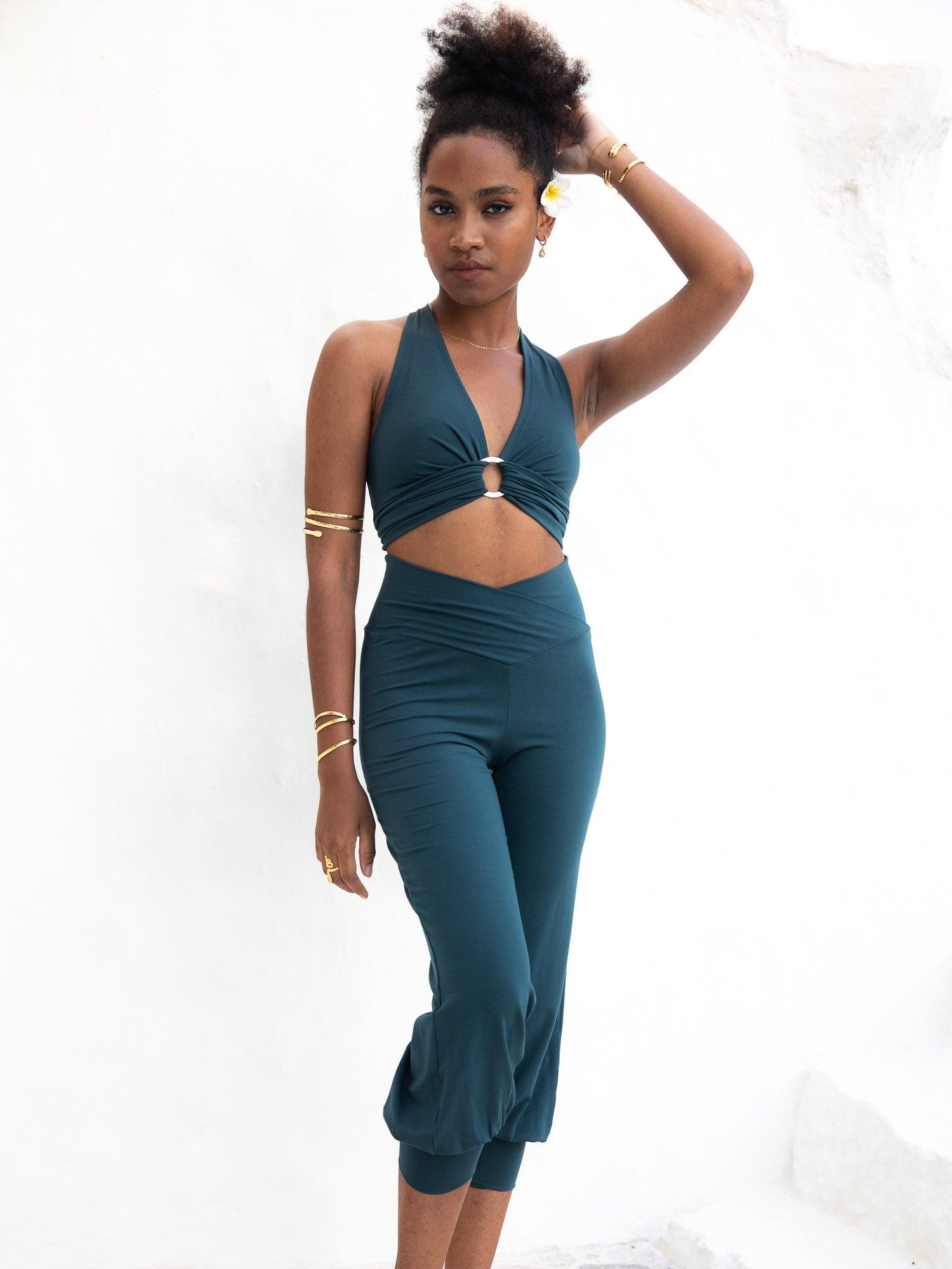 a young woman with dark skin and an afro hairstyle is modeling a tealcolored twopiece outfit. the outfit consists of a halterneck cropped top with a ring detail at the center and matching cropped capristyle pants. the pants have a gathered, slightly baggy fit at the bottom. she's accessorized with gold bracelets on her wrists. the model is posed against a plain white background, showcasing the outfit clearly. the overall aesthetic is casual, relaxed, and slightly bohemian.