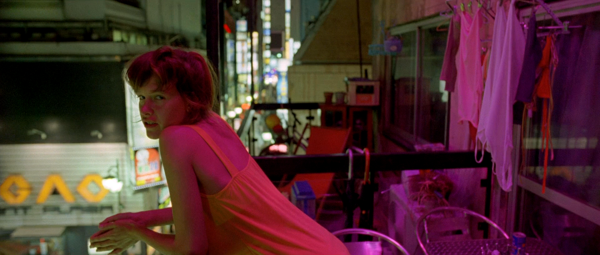 a woman with long red hair stands on a balcony, looking toward the camera with a relaxed and casual expression.  she is wearing an orange tank top and is standing in the midst of a bustling urban cityscape. the balcony is decorated with a clothesline strung with pink and white clothing and behind her, we see a lit up city with flashing neon lights. the image is bathed in a soft, warm glow.