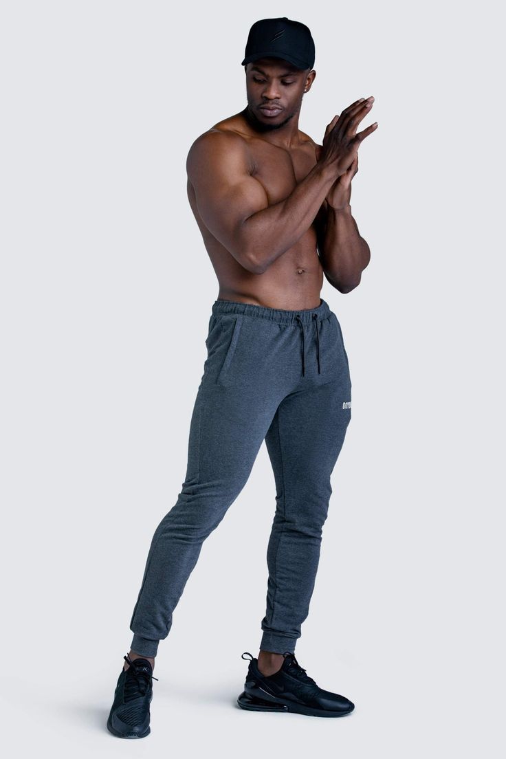 a shirtless muscular man with a black baseball cap is standing against a light grey background. he is wearing dark grey sweatpants and black air max 270 sneakers. his hands are clasped in front of him.  the overall impression is one of fitness and athleticism.