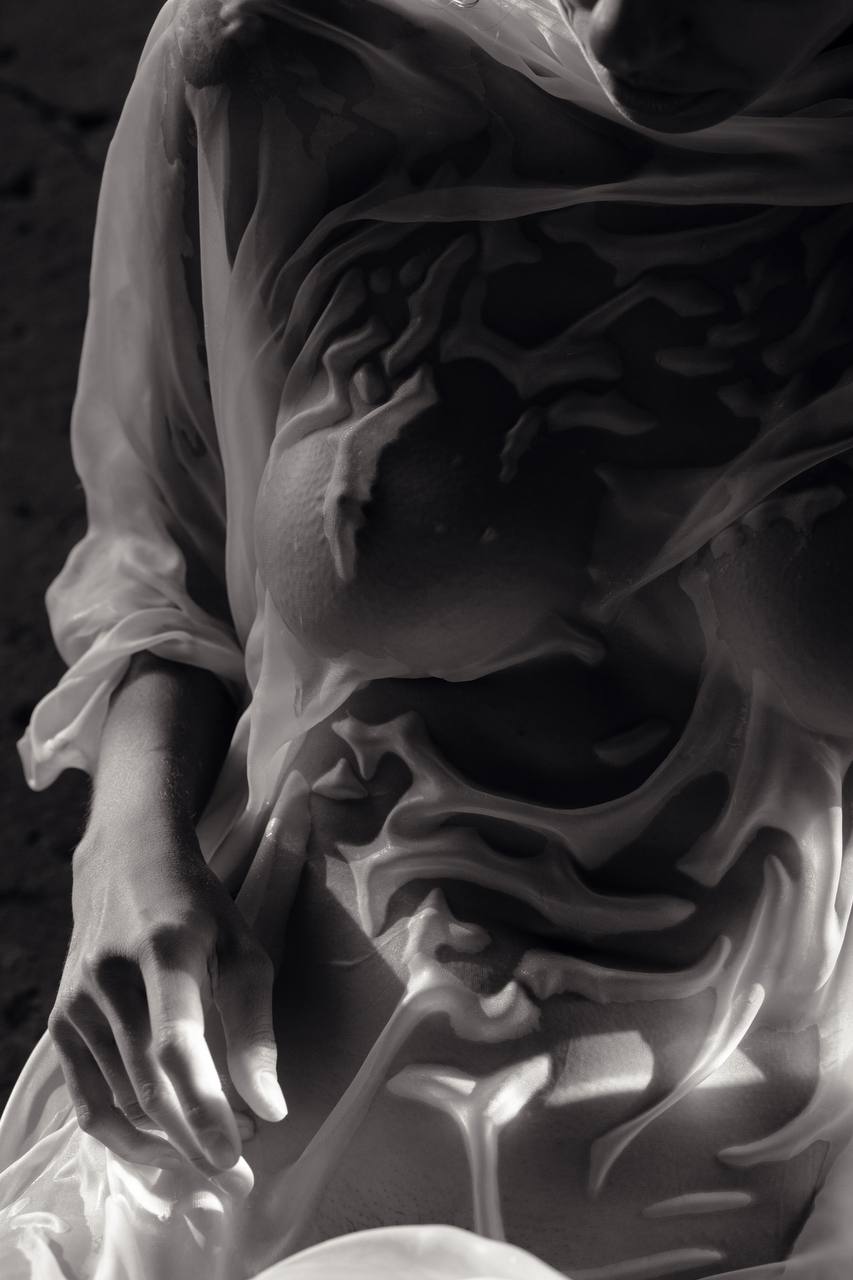 the image is a black and white, closeup photograph of a woman's torso. she is draped in a sheer, wet fabric that clings to her body, revealing its form beneath. the fabric is translucent, allowing the viewer to see the contours of her body, though not explicitly revealing any private areas. the lighting creates strong contrasts between the light and shadow, highlighting the texture of both the skin and the wet fabric. the overall feel is sensual and artistic, with an emphasis on form and texture.  the image has an abstract quality due to the way the fabric drapes and interacts with the body.