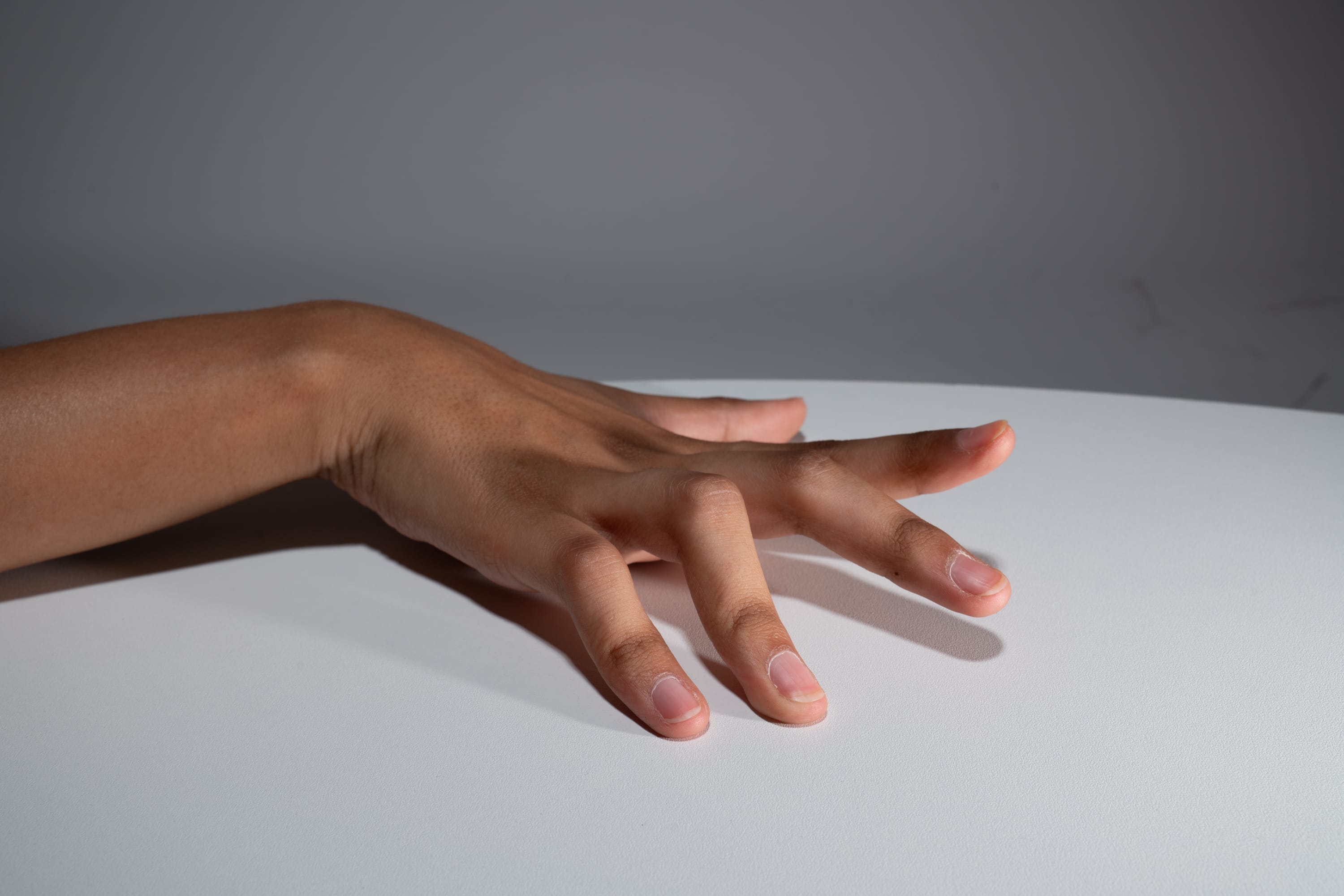 a hand with its fingers spread out is resting on a white table. the hand is illuminated by soft light, casting shadows on the surface. the background is a plain gray wall.