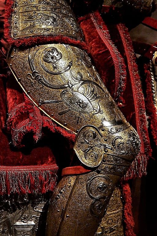 a closeup image showcasing the ornate details of a suit of armor. the armor, crafted from metal with gold accents, is adorned with intricate engravings and embellishments. it is set against a backdrop of rich, red velvet fabric, further enhancing its luxurious and historical appeal. the lighting highlights the textures and craftsmanship of both the metal and the fabric, drawing attention to the intricate patterns and the overall artistry of the piece.