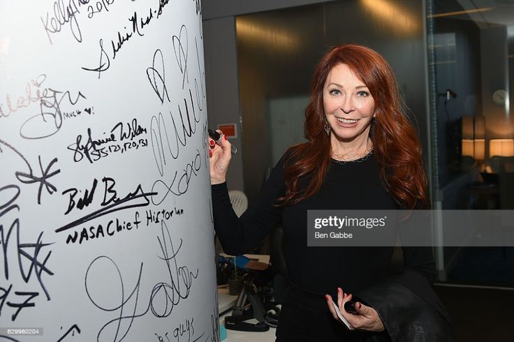 a redhaired woman in a black dress smiles as she signs an autograph on a white wall covered in signatures.