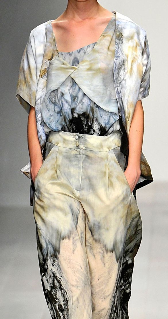 a fashion model poses in a multipiece clothing set featuring a swirling pattern. the ensemble includes highwaisted trousers, a blouse with a layered design, and a shortsleeved jacket or top. the model has their hands in the trouser pockets, showing off the waistline.