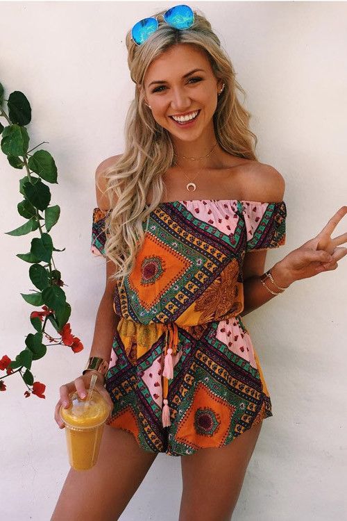 a young blonde woman is posing outdoors, wearing a colorful offtheshoulder romper and sunglasses. she's holding a glass of orange juice and flashing a peace sign, exuding a happy and carefree summer vibe.
