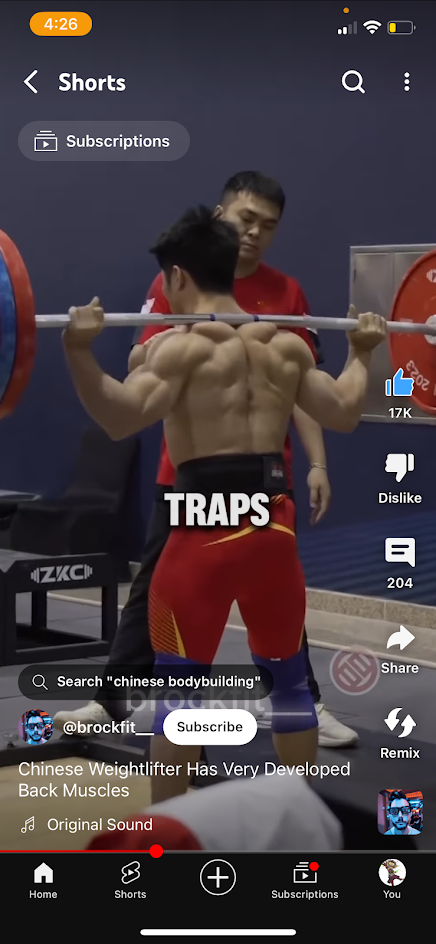 a man in a red and yellow shorts is doing a weightlifting exercise. he is lifting a barbell over his head and his back muscles are very defined. the text on the screen says "traps", indicating that the exercise is targeting the trapezius muscle. the video is from an app called "shorts" and the user's name is @brockfit.  the title of the video is "chinese weightlifter has very developed back muscles". the video is tagged with "chinese bodybuilding".