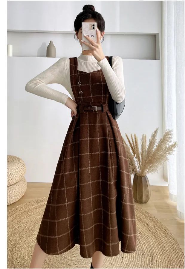 a woman is standing indoors, modeling a brown and white plaid jumper dress. she is also wearing a white turtleneck underneath the dress. the dress has a belted waist and button detailing on the bodice. the image is welllit, showcasing the details and texture of the outfit.