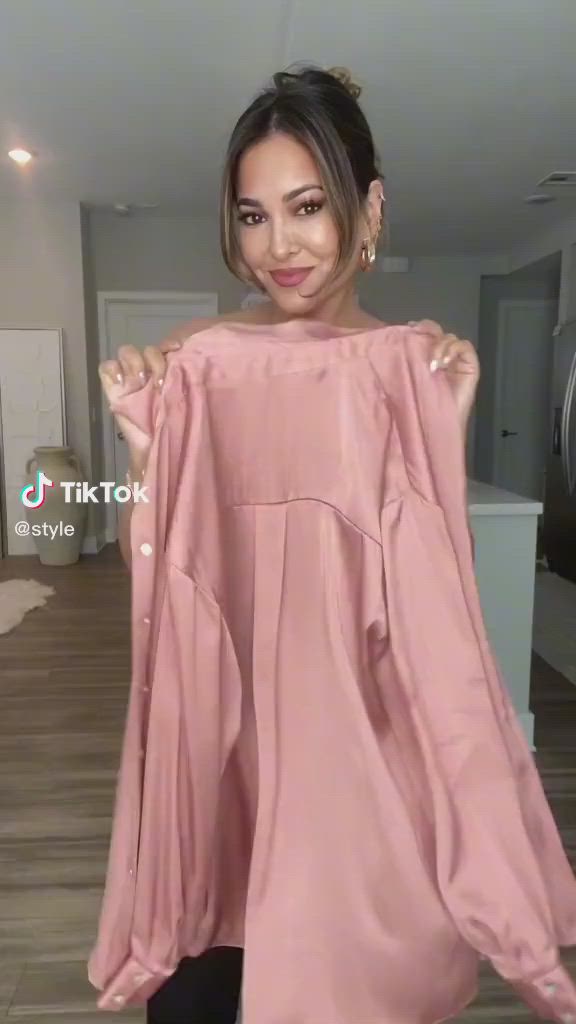 a woman is holding a pink long sleeve shirt in front of her. the video is from tiktok and the creator's handle is @style. she is standing indoors with neutralcolored walls. the woman has a hairstyle with some of her hair pulled back. she is wearing earrings.