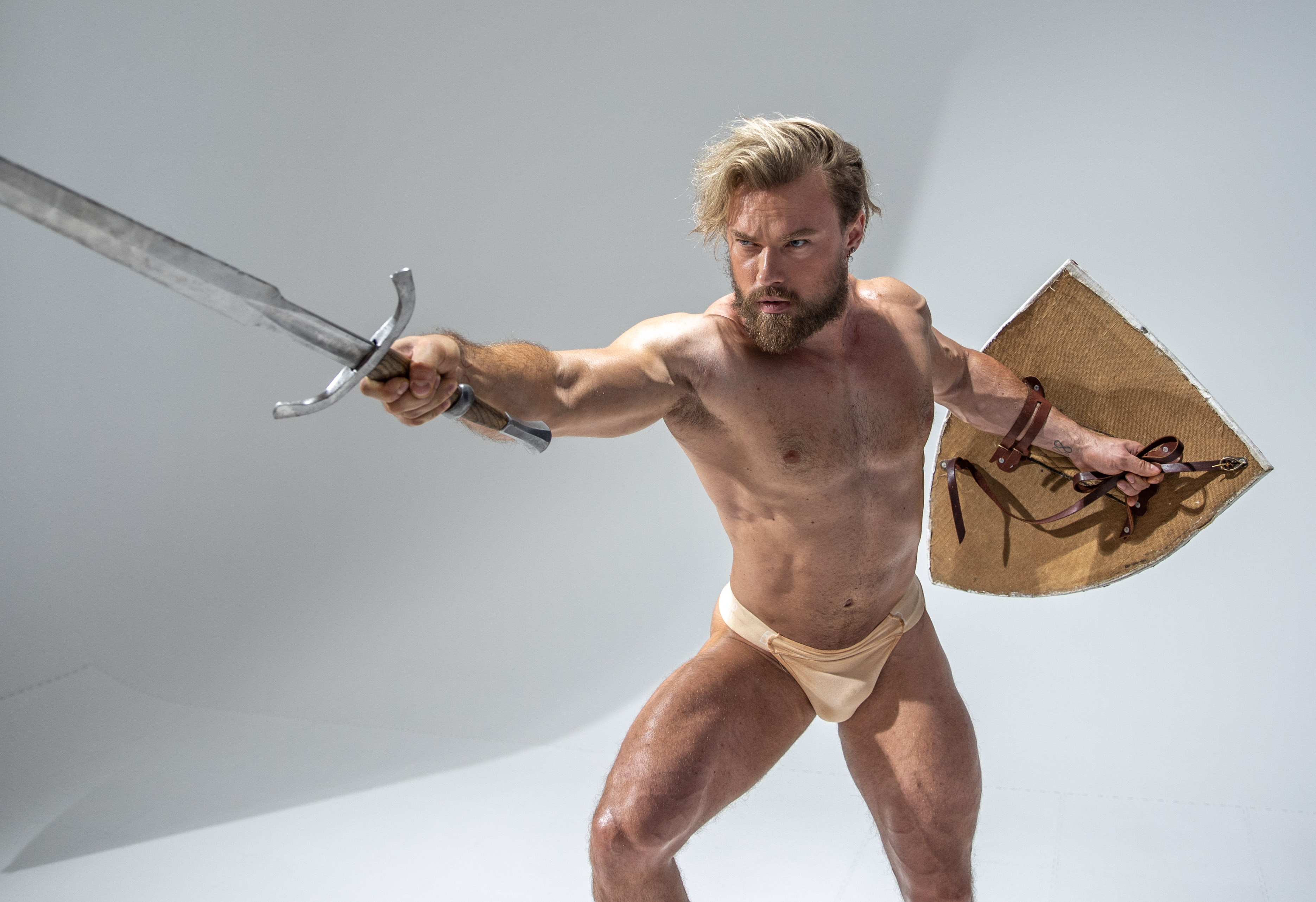 a muscular man with a beard and blonde hair, wearing only a loincloth, stands in an attack pose against a white background. he holds a sword in his right hand and a round shield in his left. the image emphasizes his physique and the power of his stance. the studio lighting highlights his muscles and the metallic glint of the sword.