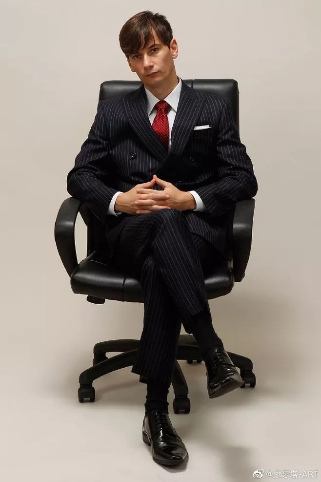 a man in a black pinstripe suit and red tie sits confidently in an office chair. he is looking directly at the camera with a serious expression.