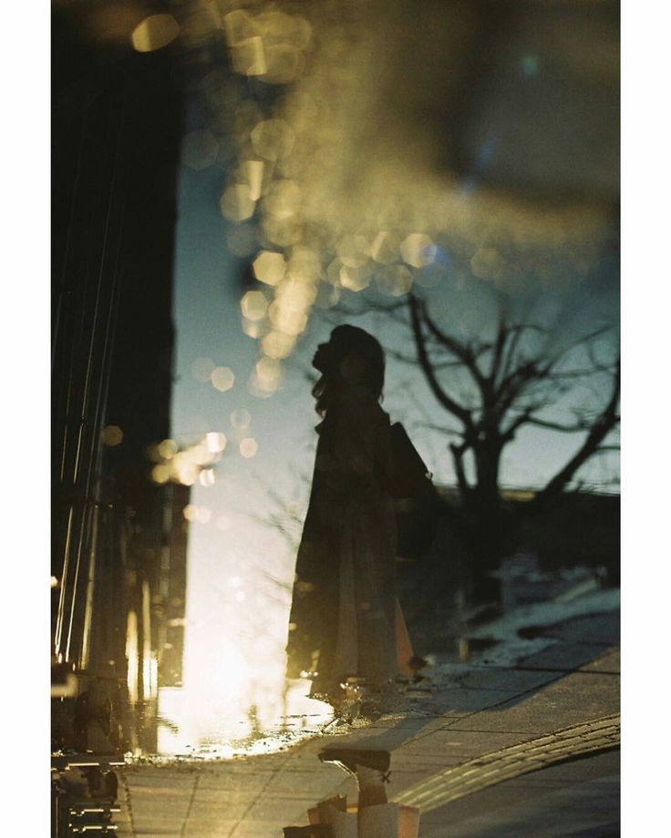 the image is a moody, atmospheric photograph taken with film. it features the silhouette of a woman in a long coat standing near a puddle on a city street. the puddle reflects the setting sun, creating a golden hour effect and bokeh.  the reflection also shows a portion of the surrounding urban environment, including trees and buildings.  the overall mood is contemplative and slightly melancholic, enhanced by the use of shadow, low light, and the woman's solitary figure. the photography style is reminiscent of analog film, with light leaks and a slightly grainy quality. the image evokes a sense of mystery and quiet solitude.