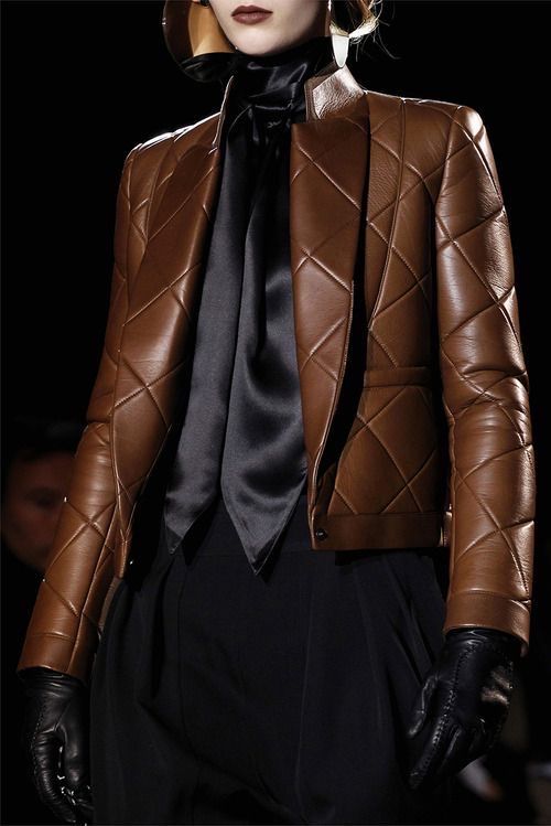 a woman model walks the runway wearing a brown leather jacket and black gloves. the jacket is quilted and has a black satin tie at the neckline. the model is wearing black pants and has a serious expression on her face. she is showcasing a fall/winter fashion trend.