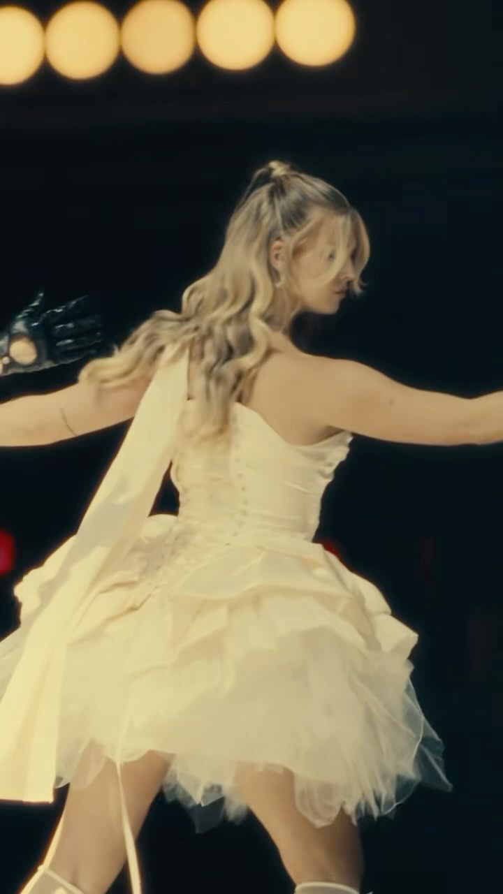 a woman with blonde hair in a high ponytail wearing a short, oneshoulder white dress with a tulle skirt is on stage. she is wearing black gloves on one hand. the stage has round lights in the background. she appears to be dancing or performing.