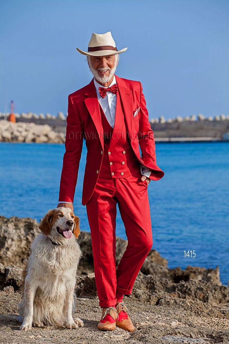 an older, stylish man in a vibrant red threepiece suit and straw cowboy hat stands by the ocean with a white and brown dog. the man is smiling and the dog is sitting calmly.  the scene is set against a rocky coast with blue water in the background. the image is welllit and professional, possibly for a fashion catalog or magazine. a watermark with "ottavionuccio.com" and the number "1415" is visible on the image.