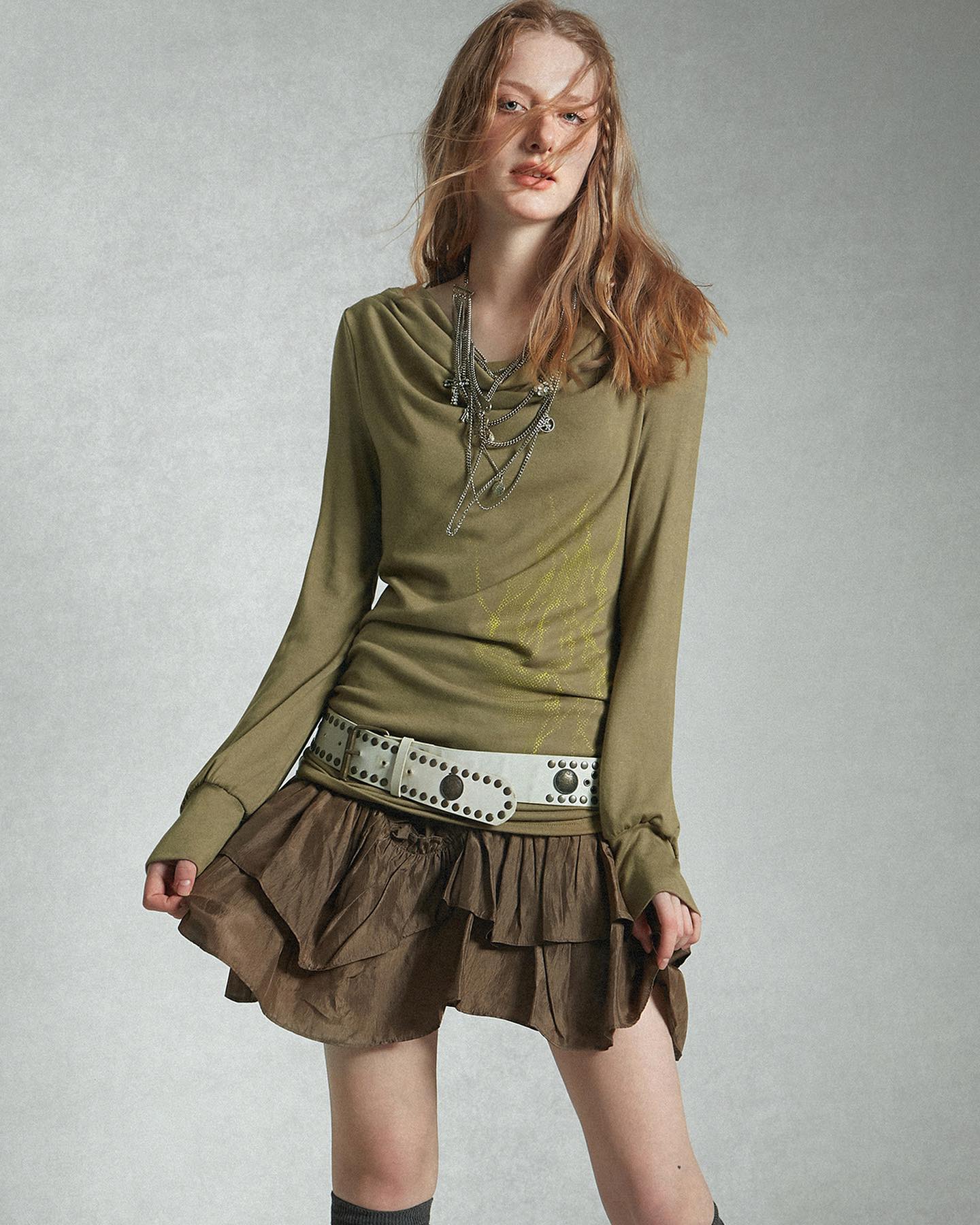 a fullbody shot of a young woman model posing against a light grey background. she's wearing a longsleeved olive green cowlneck top, a brown tiered ruffle skirt, a studded white belt, and a layered silver necklace. her blonde hair is styled in loose braids. the overall style is casual, bohemian, and slightly vintage. the image is welllit and professionally photographed.