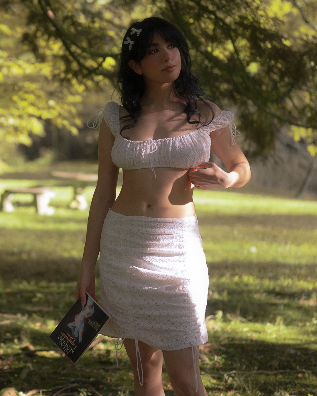 a woman stands outdoors in a grassy park or forest setting. she is wearing a white twopiece outfit consisting of a lace skirt and a matching top. the woman has dark brunette hair with bangs, and she is wearing a hair clip. she is holding a book in her hand. the scene is illuminated by sunlight filtering through the trees. there is a bench in the background.