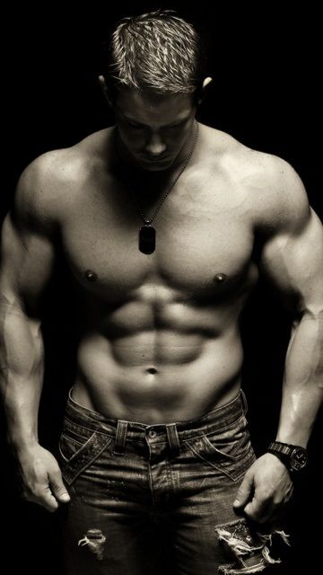 a muscular man is shirtless, showing off his toned physique. he is wearing jeans and stands with a pensive expression in a dark studio. the photo is in black and white.