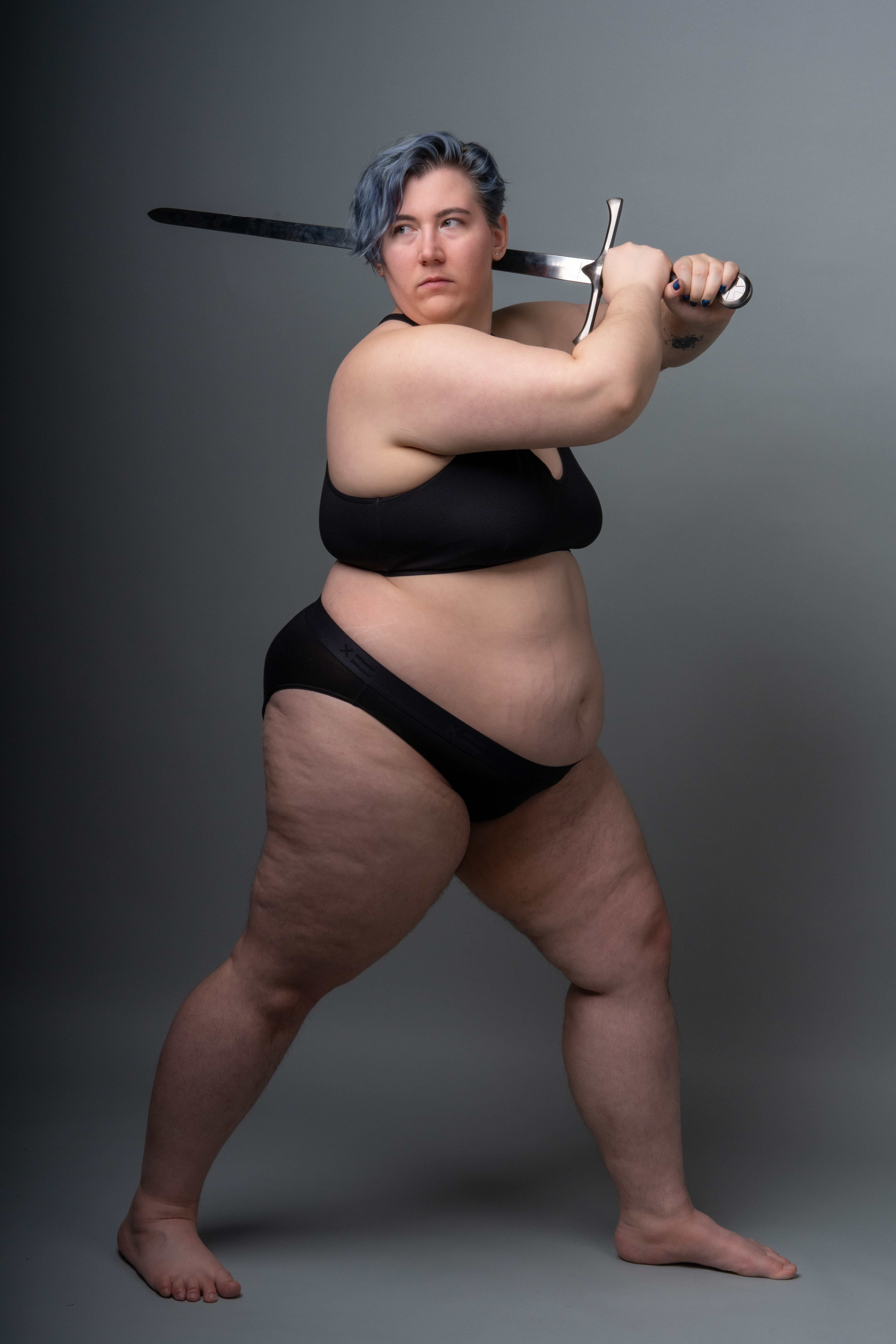 a full body studio shot of a woman with blue hair holding a sword in a defensive stance. she is wearing a black bra and black underwear against a gray background.