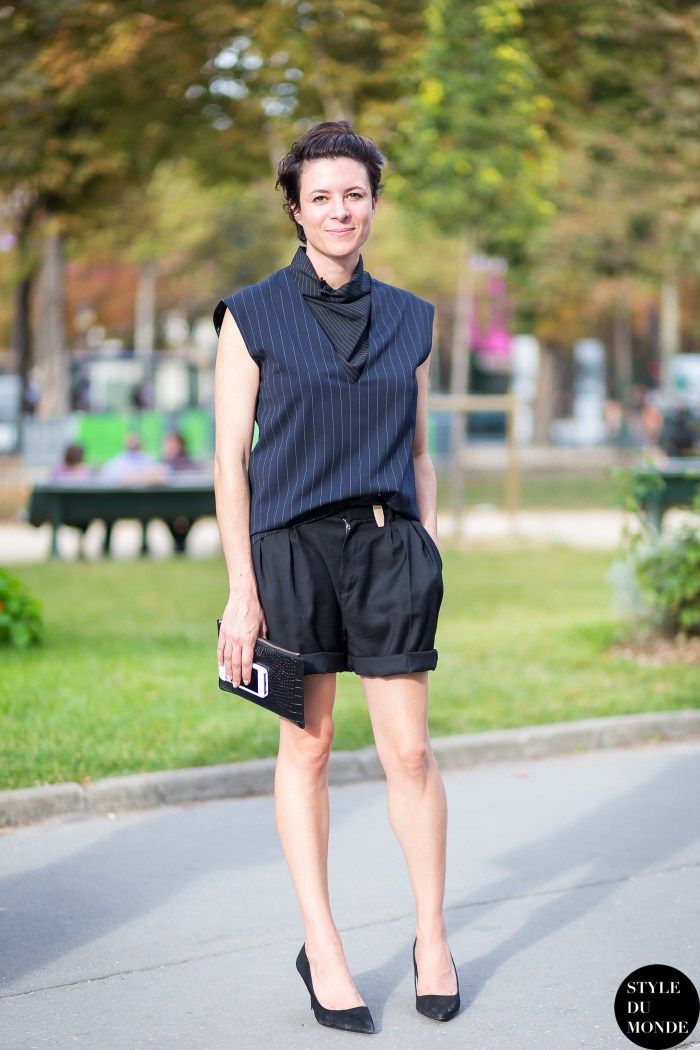 a woman is standing outdoors in a stylish outfit consisting of a pinstripe sleeveless top with a black neck scarf, black cuffed shorts, and black heels. she is holding a clutch. the background features trees and greenery, suggesting an urban park or street setting.