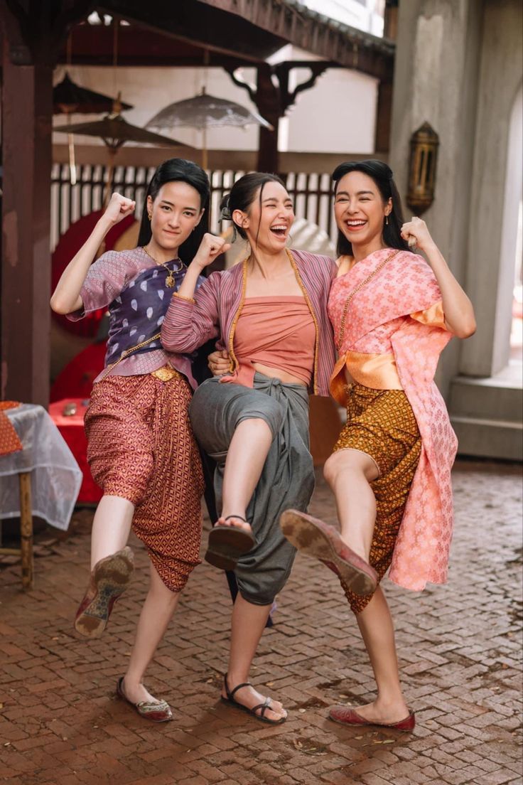 three women are laughing and kicking their feet up in the air. they are wearing traditional thai clothing. they are standing on a brick floor.
