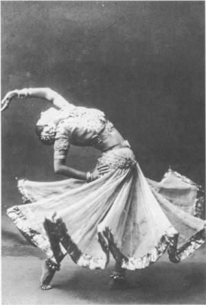 a black and white photo shows a dancer in midperformance, bending backwards in a classical indian dance pose. she is wearing a traditional skirt that is flared out, along with jewelry. the photo is a historical image.