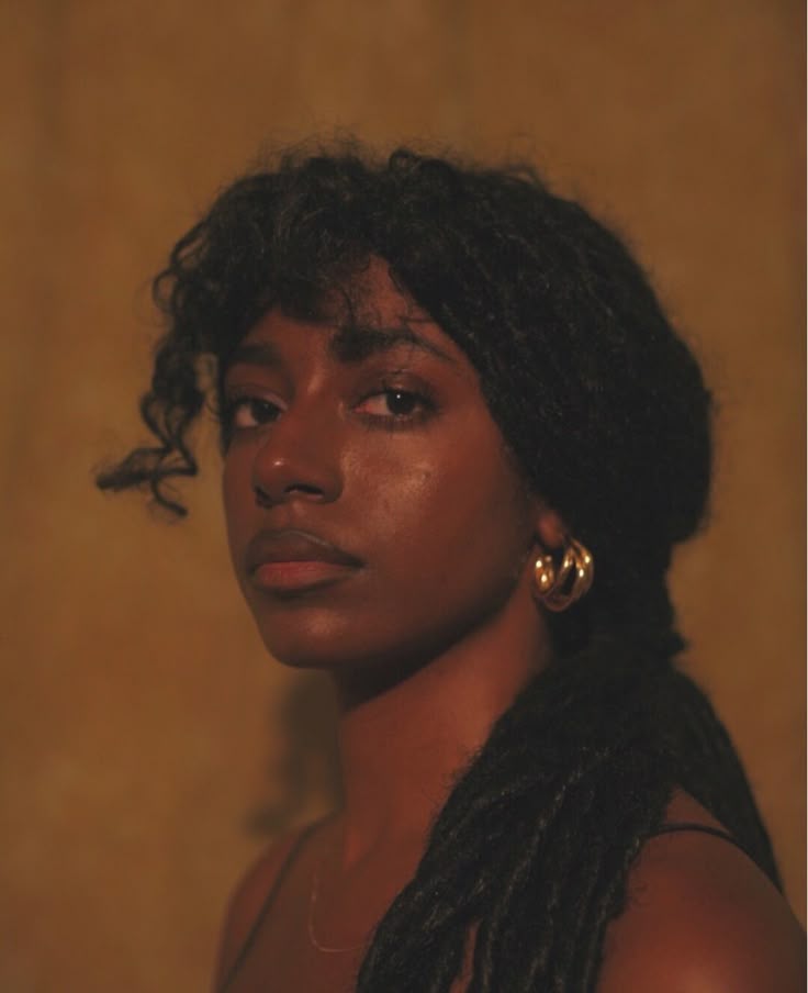 a portrait of a woman with dark skin and black hair wearing gold earrings, looking directly at the camera with a calm and pensive expression. she has a subtle necklace on. the lighting creates a warm and intimate atmosphere.