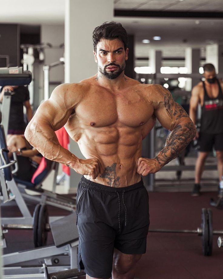 a muscular man in black shorts, wearing a tattoo on his arm, is standing in a gym. he is looking at the camera with a serious expression. he is posing with his arms slightly bent, displaying his bicep, tricep and abdominal muscles. the gym is wellequipped with weights and other equipment.