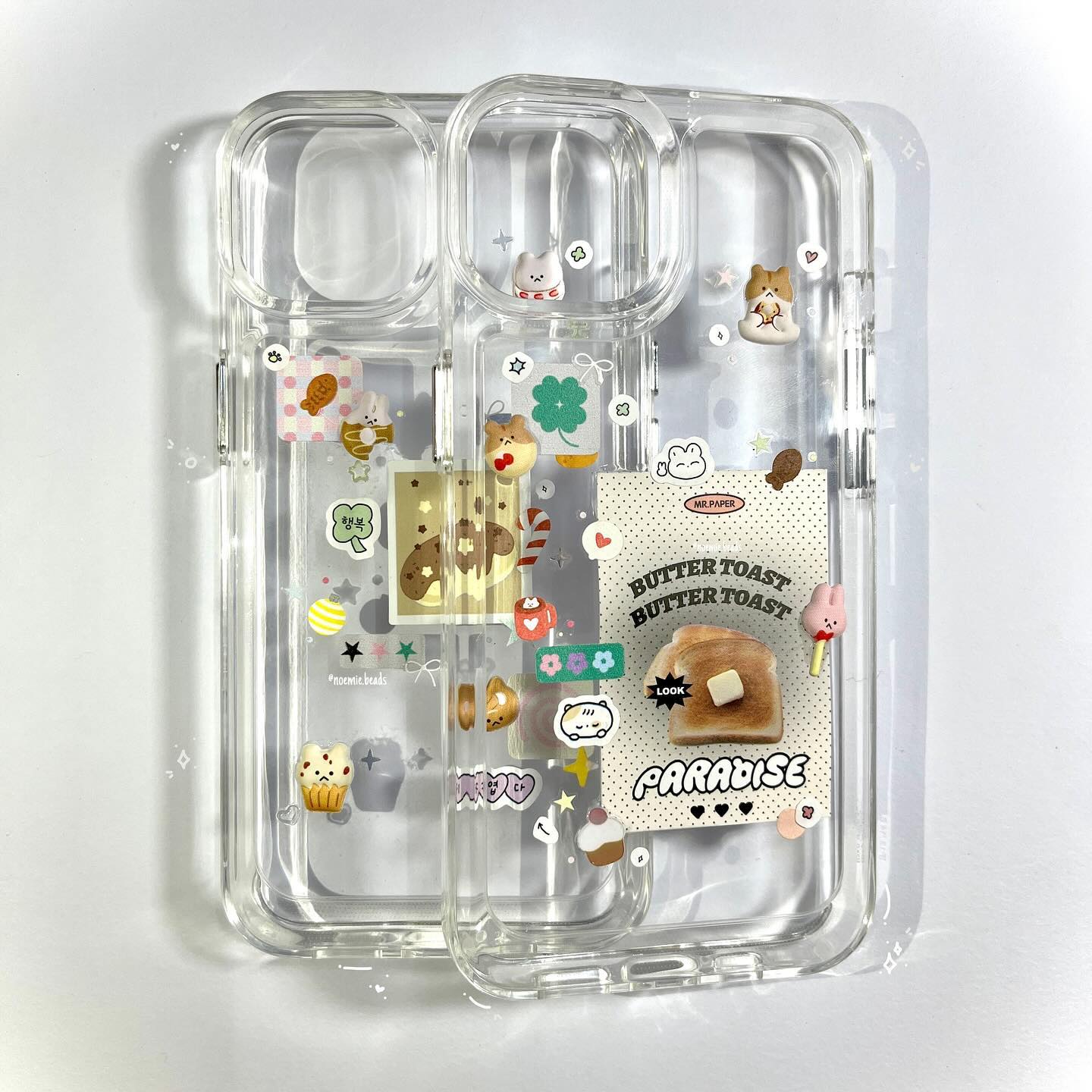 a clear phone case decorated with various stickers, including images of buttered toast, a bear, a clover, and a cupcake, against a white background.