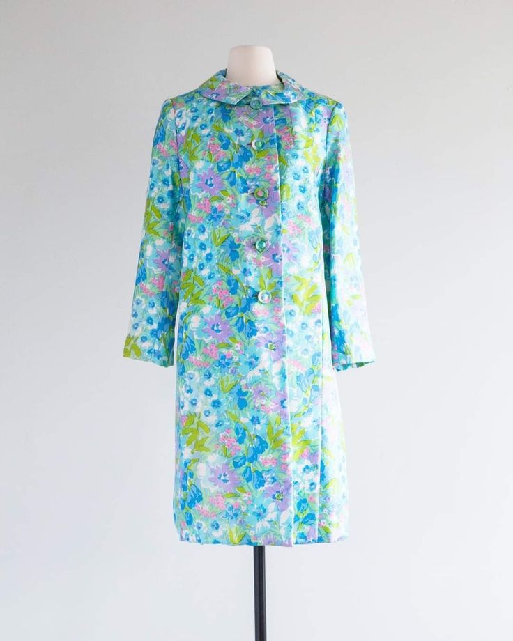 a floralprint coat is displayed on a mannequin against a white backdrop. the coat features a pattern with blue, green, and purple floral designs. it has long sleeves, a peter pan collar, and a buttondown closure.