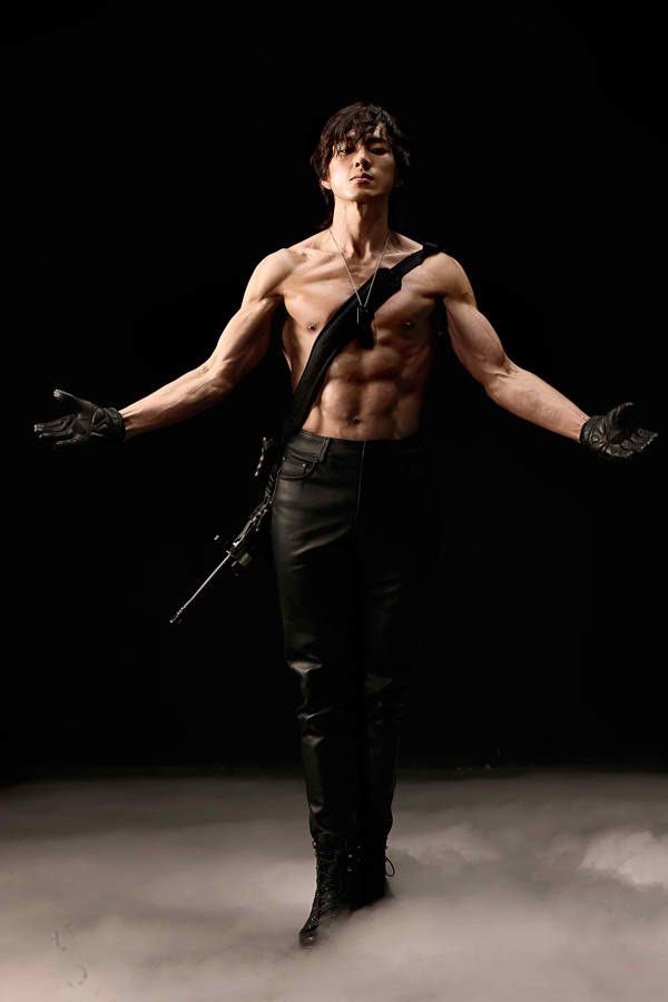 a shirtless man with a muscular physique is posing with his arms outstretched in a dark studio. he's wearing black leather pants, black boots, and black gloves. there's a hint of fog or smoke in the air, adding to the dramatic and mysterious atmosphere.