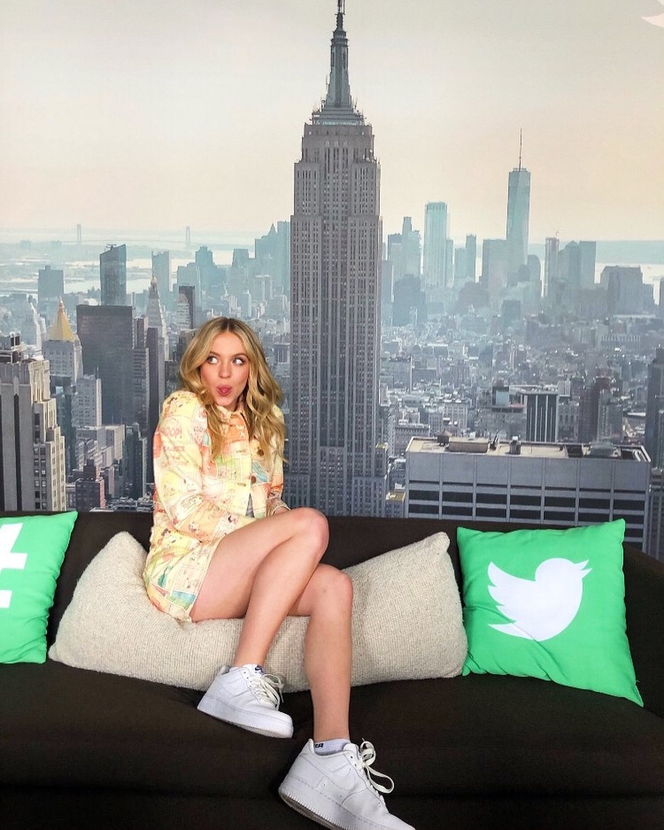 a blonde woman in a pastelcolored dress sits playfully on a darkcolored couch.  she's wearing white sneakers and is posing for a photo. behind her is a large backdrop featuring a scenic view of the new york city skyline, including the empire state building.  the couch has green pillows with the twitter logo on them, suggesting a social mediarelated photoshoot. the overall vibe is lighthearted and stylish.