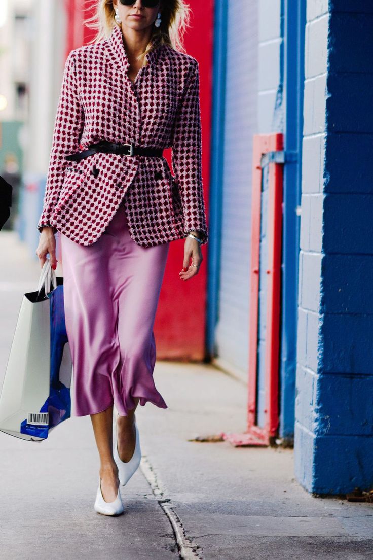 a stylish woman strolls down a city street, her outfit a blend of sophisticated and casual chic. she wears a pink slip dress paired with a red and white checkered blazer, cinched at the waist with a black belt.  white pointedtoe heels complete the look, adding a touch of elegance. the woman carries shopping bags, suggesting a day of shopping. the vibrant backdrop of red and blue painted walls provides a visually striking contrast to her outfit.