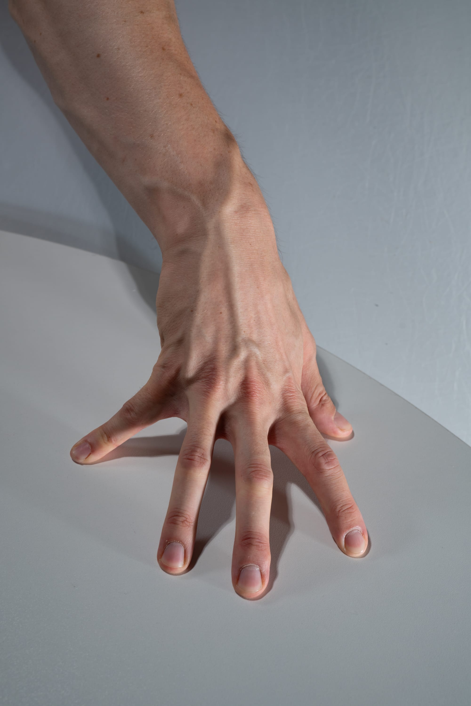 a man's left hand and forearm are resting on a white surface. the hand is spread out, with the fingers slightly curled. the veins in the hand and arm are visible. the image is brightly lit and taken from a topdown perspective.