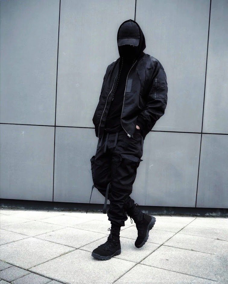 a man in all black clothing is standing in front of a gray wall, his face is hidden by a black cap and his hands are in his pockets. the man's clothing includes a bomber jacket, black pants, and black boots.