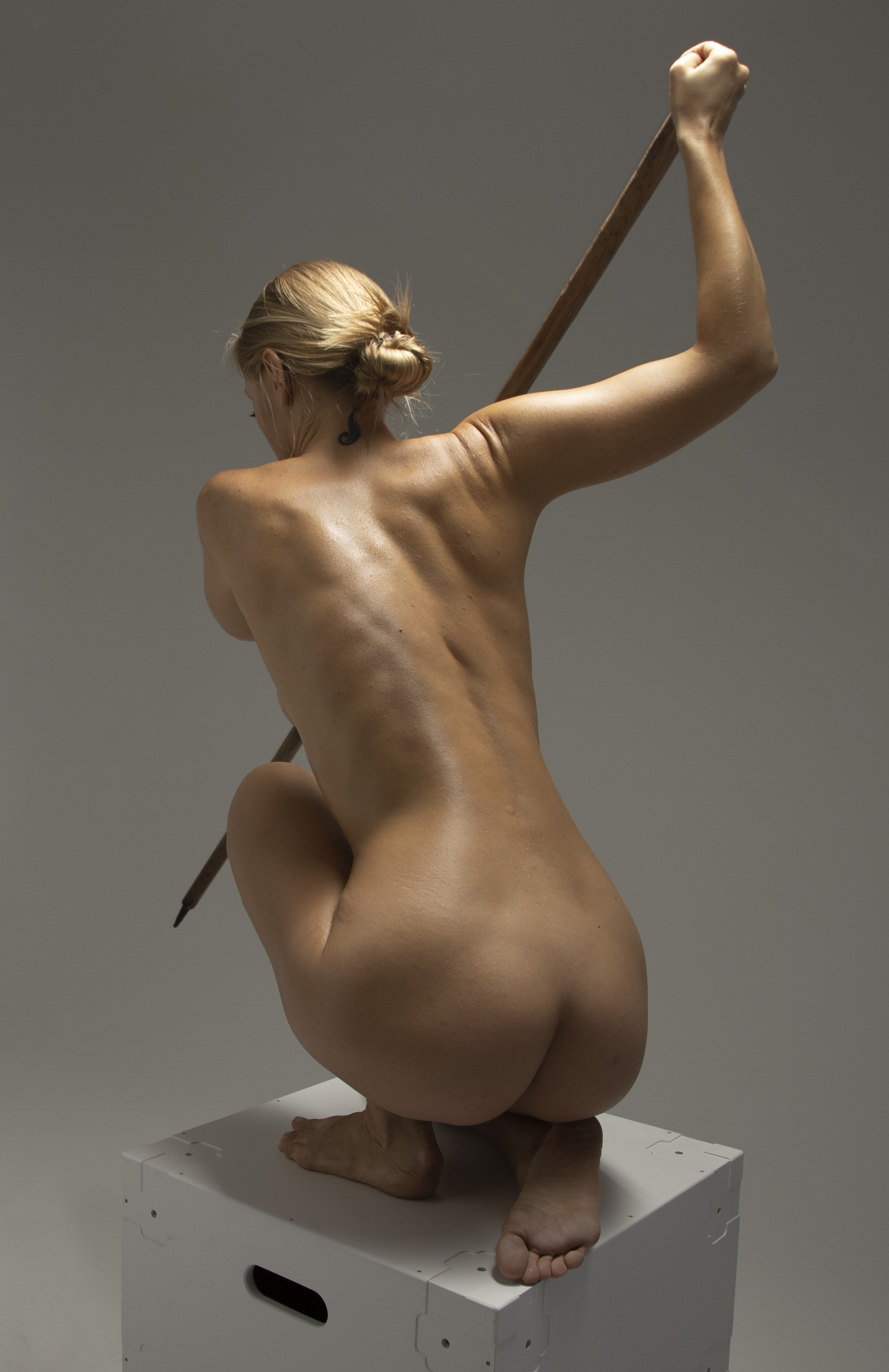 a nude woman is crouching on top of a white box, facing away from the camera. she is holding a stick with both hands, her right arm raised in the air. her blonde hair is in a bun, and there is a tattoo on her neck. she is barefoot.
