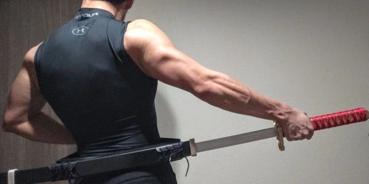 the image shows the back of a muscular man wearing a black under armour sleeveless shirt. he is holding a katana (a japanese sword) with a redwrapped hilt, drawing it from a black leather scabbard attached to his waist. his arms are visible, showcasing prominent musculature and veins. the background is a plain, light gray wall. the overall impression is one of strength and martial arts practice or training.