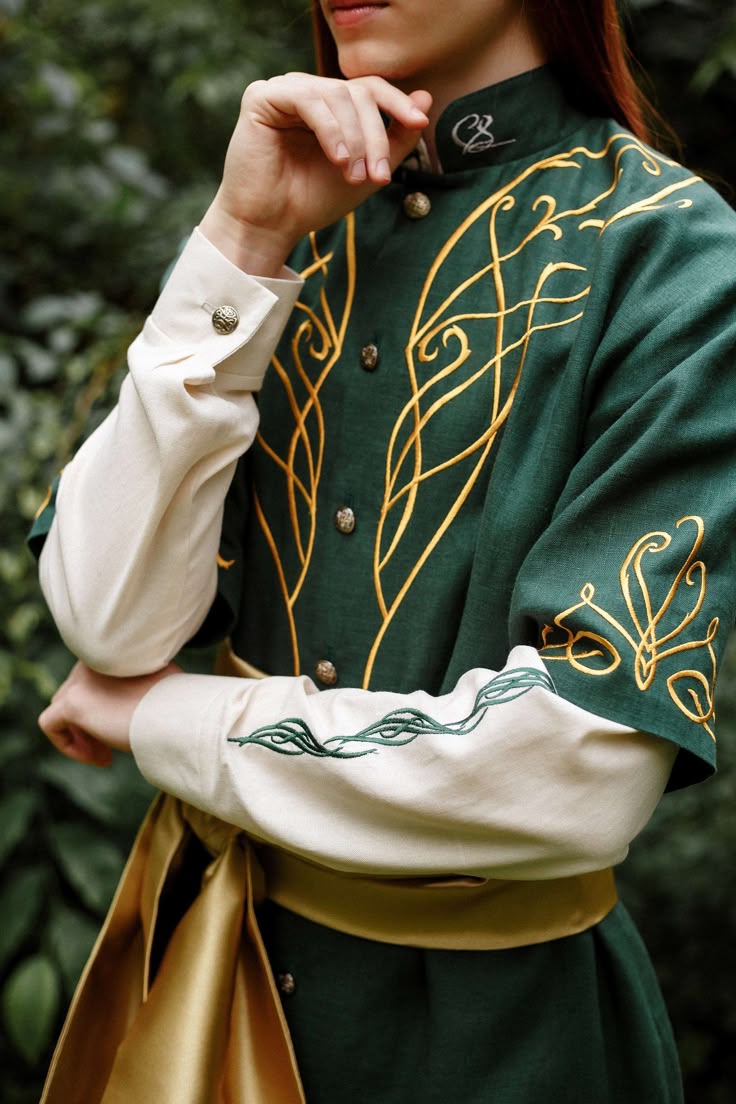 a person in an elf costume is standing in front of some green foliage. they are wearing a green shirt with gold embroidery and white cuffs with a small green vine embroidered on each cuff. the shirt has small gold buttons. a yellow sash is tied around their waist. the person's hand is up near their face, and the rest of their body is cut off in the image.