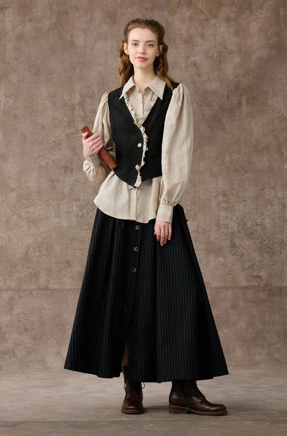 a young woman with long brown hair wearing a white blouse, black vest, black pinstripe skirt and brown boots stands against a plain brown background. she is looking directly at the camera.