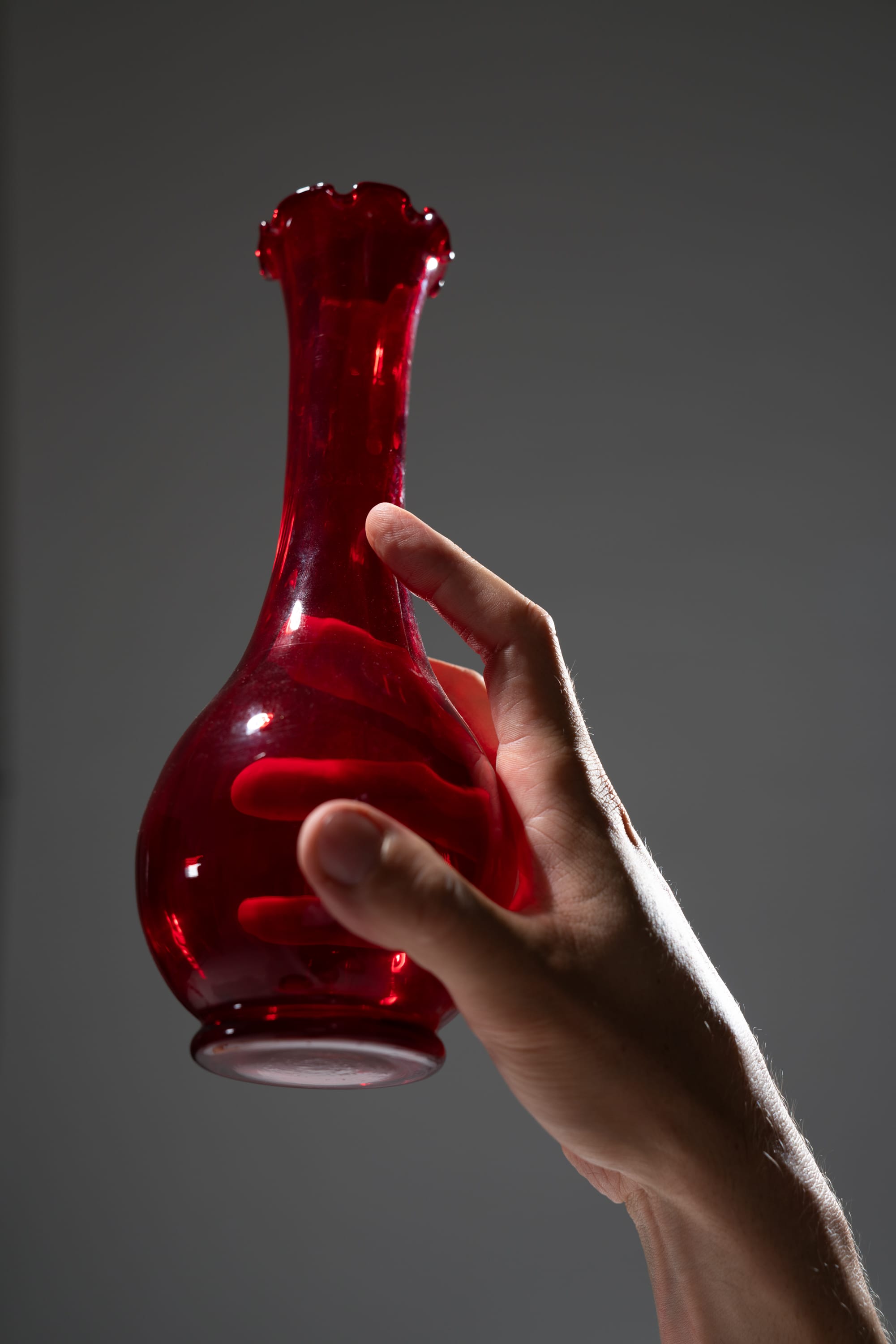 a hand gracefully holds a red glass vase against a muted grey background. the studio lighting illuminates the vase and hand, highlighting their elegant form.