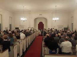 an indoor view of a church during a wedding ceremony. people are gathered in pews on both sides of a red carpet that leads to the altar. chandeliers hang from the ceiling, illuminating the scene. the church is filled with attendees participating in the event.