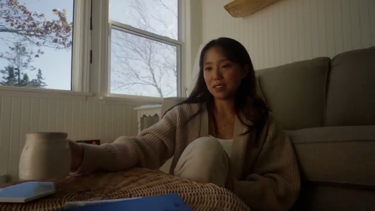 a woman is sitting on the floor in a living room, drinking from a mug. she is wearing a beige cardigan and white pants. there is a window behind her with a view of trees. the lighting is soft and natural. the overall atmosphere is cozy and relaxed.