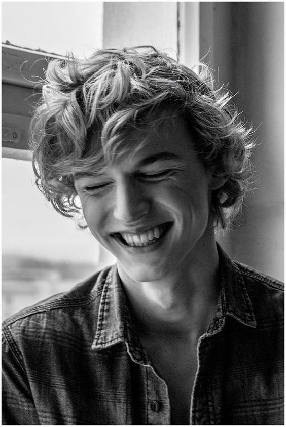 a young man with curly hair is smiling happily. he is wearing a casual denim shirt and looking down. the image is in black and white.