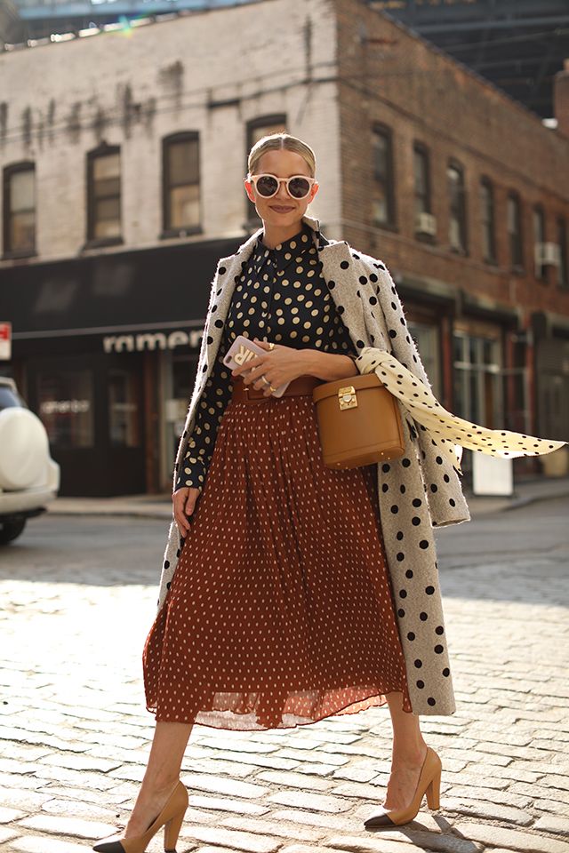 a stylish woman strolls down a city street, her outfit a chic blend of autumnal colors and patterns. she wears a rustcolored pleated midi skirt and a black and white polka dot blouse, layered under a grey polka dot coat.  beige heeled shoes and a brown tophandle bag complete the look.  she's accessorized with round sunglasses. the backdrop is a classic city street with brick pavement and brownstone buildings. the overall image is one of sophisticated and effortless style.