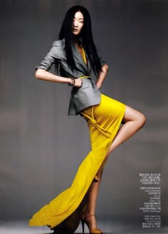 a woman wearing a grey blazer and a yellow skirt poses for a studio shoot.  the woman has long black hair and is wearing high heels.  she looks to be in a casual, relaxed pose.  she is looking to the side and has a slight smile on her face.