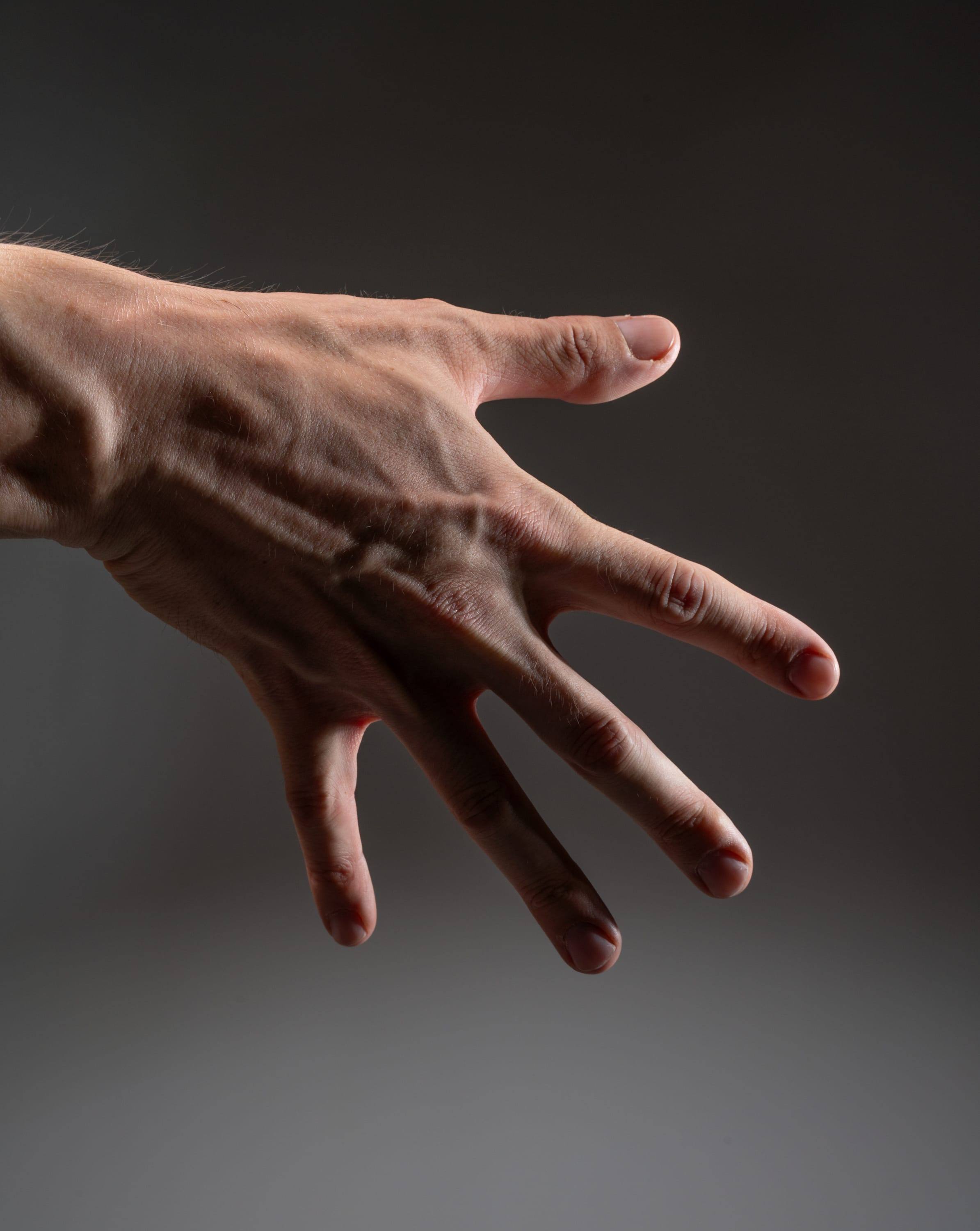 a closeup shot of a man's outstretched hand against a dark gray background. the image emphasizes the contours of the hand, the prominent veins, and the texture of the skin. the hand is illuminated by dramatic side lighting that casts deep shadows, creating a sense of depth and highlighting the details of the hand.