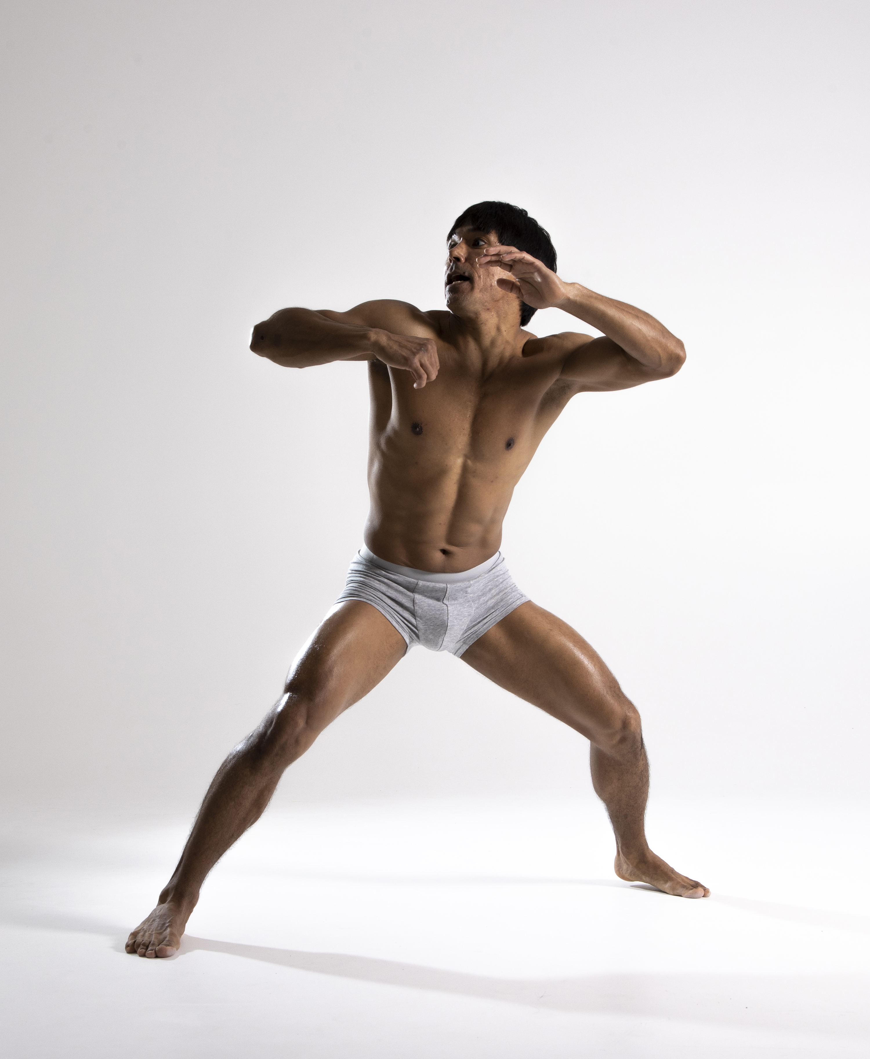 a shirtless, muscular man in grey underwear is posed dynamically against a white background. he appears to be dancing or dodging, with his hands up defensively. his legs are wide apart, and his body is oiled. the photo is a full body shot, capturing the dramatic tension of the pose.