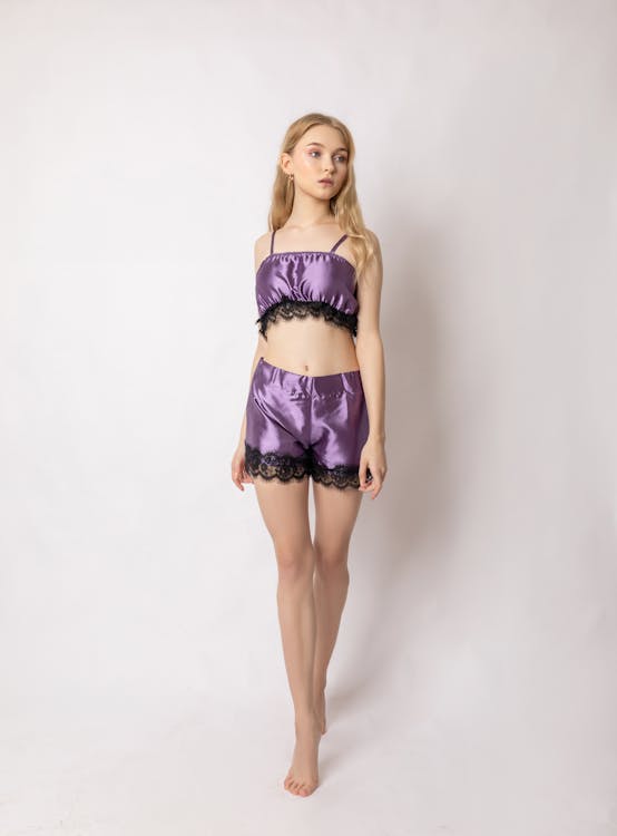 a fulllength studio shot of a young blonde woman wearing a purple satin lingerie set. the set consists of a cropped top and shorts, both trimmed with black lace. the woman is standing barefoot, her body facing slightly towards the camera. the background is plain white. the overall impression is one of elegance and sensuality.