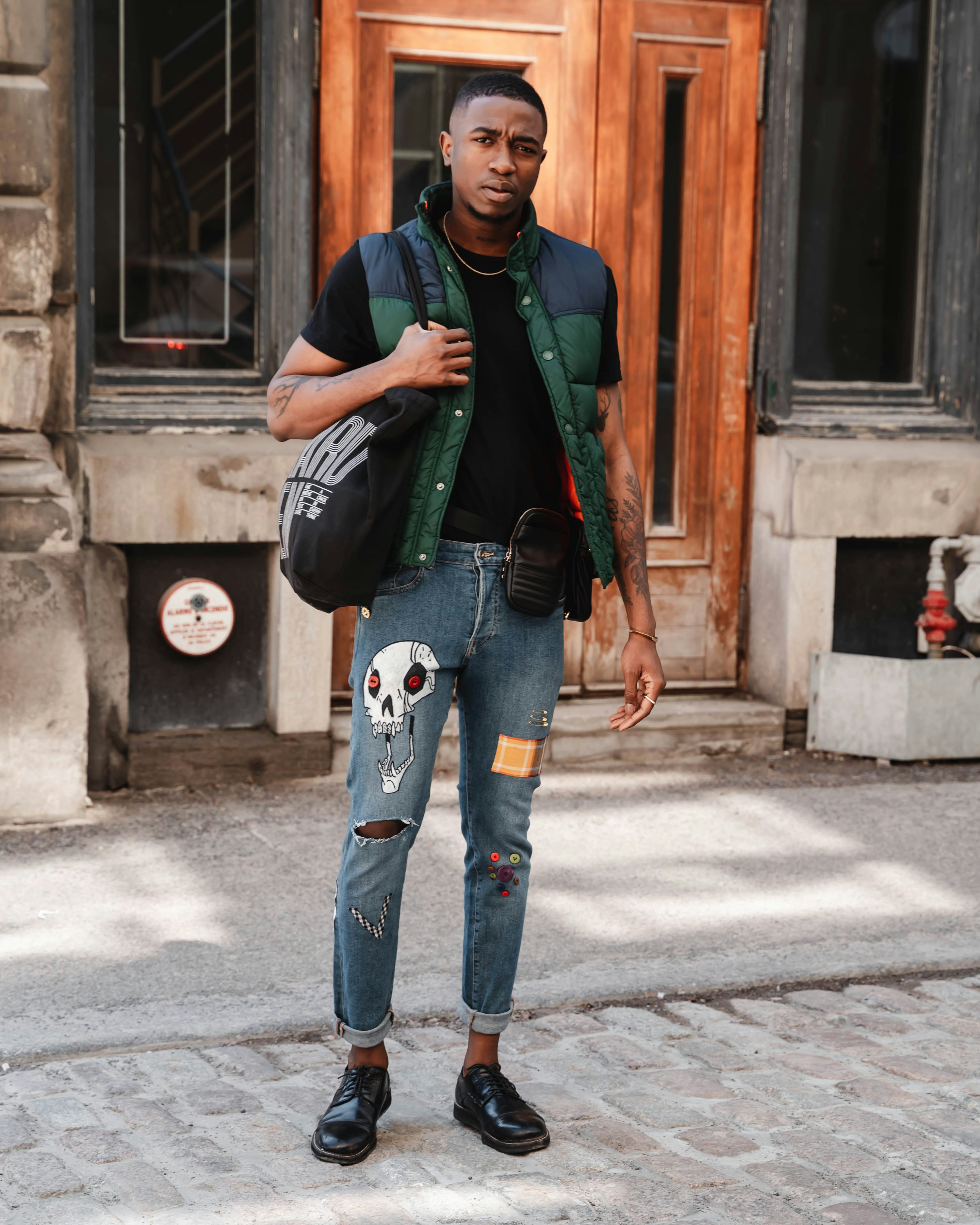 a stylish young black man stands on a city street, posing for a fashion photograph.  he's wearing a dark green and black puffer vest over a black tshirt, uniquely patched jeans featuring a skull graphic, and black oxford shoes. he carries a black tote bag and a small crossbody bag.  the background shows a classic city building with a wooden door. the overall style is urban and trendy, highlighting the model's individual fashion choices.